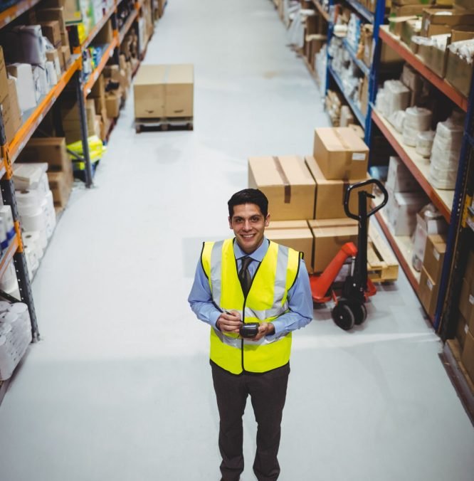 Male warehouse worker in safety vest scanning boxes in warehouse aisle with scanner and pallet jack. Industrial, logistics, organization, inventory, distribution, transportation, efficiency