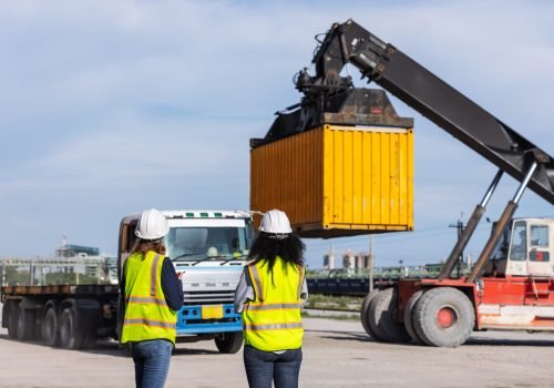 Two female workers in safety vests and hard hats oversee container loading operations at a shipping yard. A crane lifts a yellow container onto a truck, highlighting teamwork and global logistics.