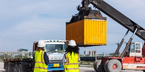 Two female workers in safety vests and hard hats oversee container loading operations at a shipping yard. A crane lifts a yellow container onto a truck, highlighting teamwork and global logistics.